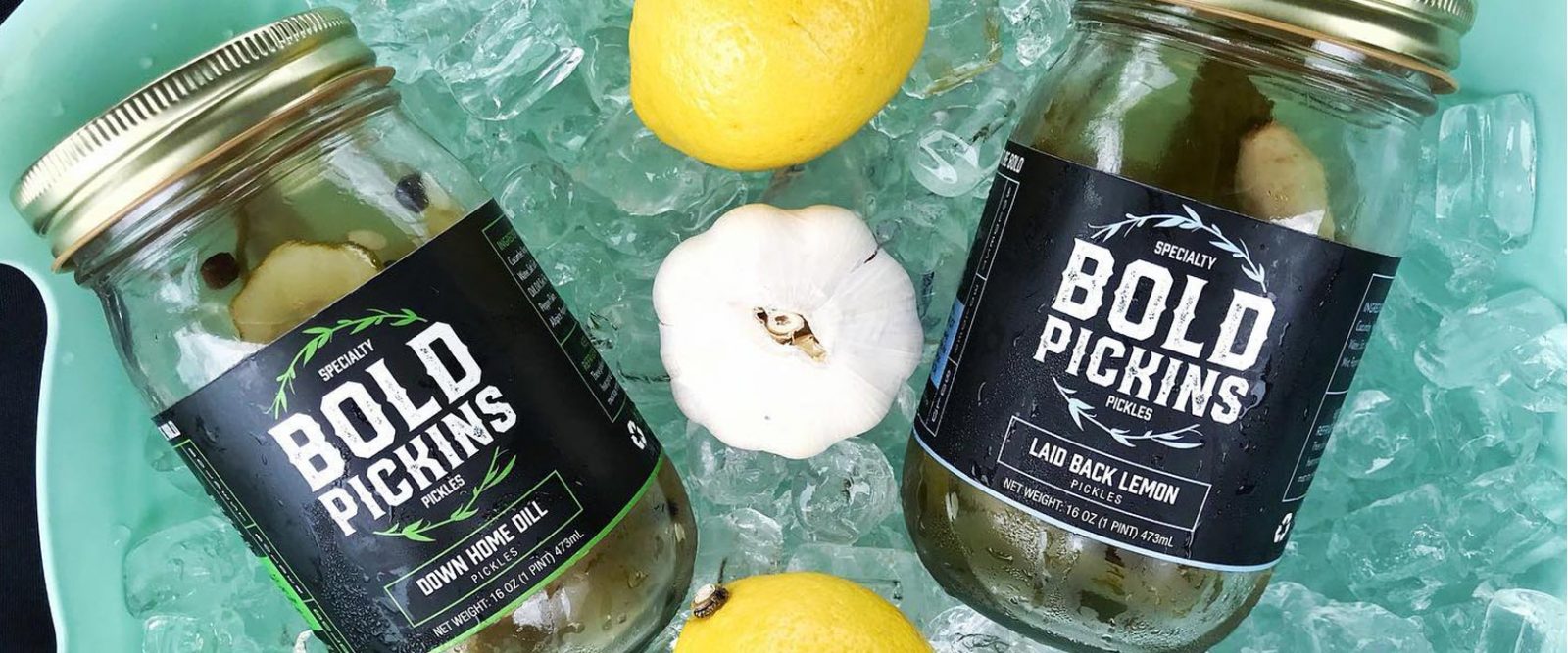 Two jars of Bold Pickin’s refrigerator pickles rising on a bed of ice with a head of garlic and two lemons