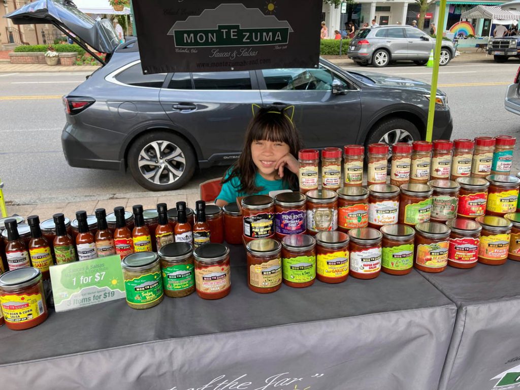 Girl seated at outdoor market table behind display of Chuck Evans’ Montezuma Sauces