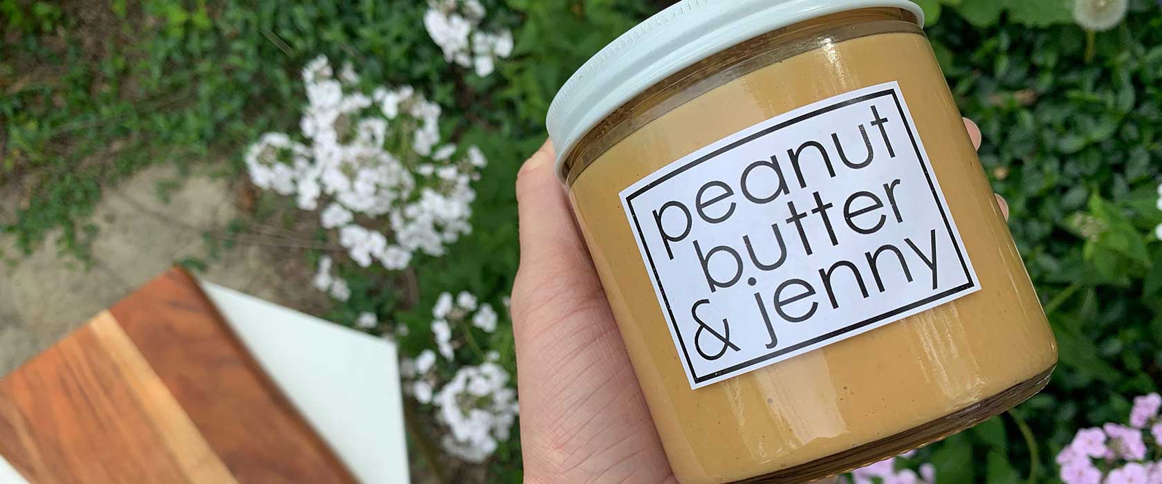 Woman's hand holding glass jar of peanut butter with label reading Peanut Butter & Jenny