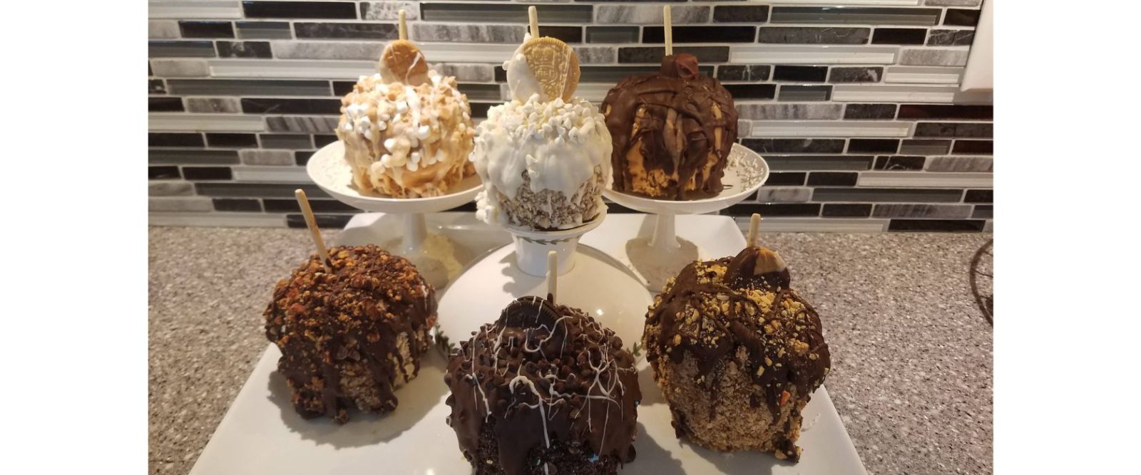 6 caramel apples, coated in different flavors of candy and toppings on white pedestals in front of black and white tiled wall