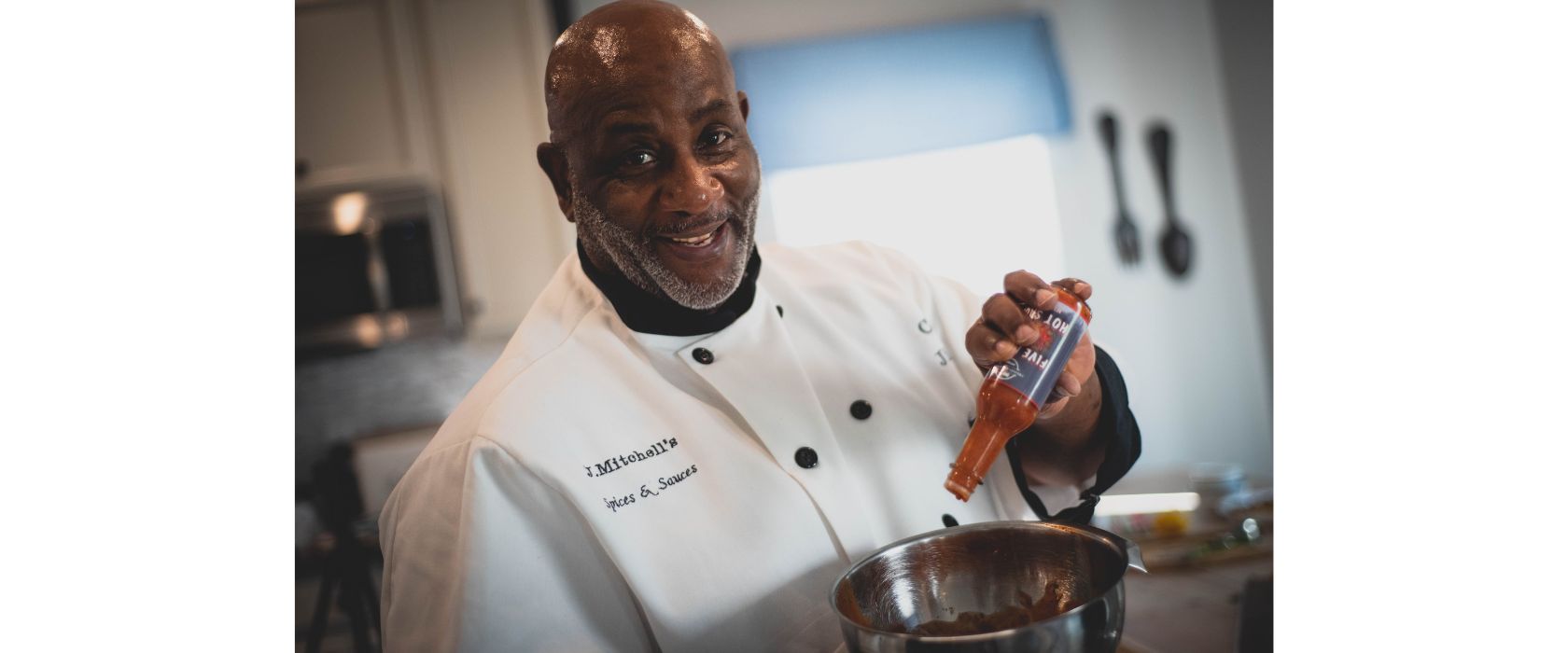 Smiling man in a white chef's coat pouring hot sauce into a metal bowl