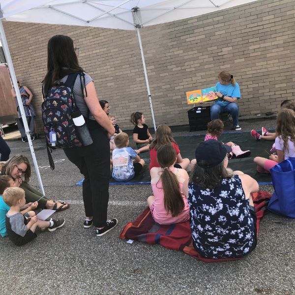 kids sitting outdoors in a parking lot under a pop up tent watching a librarian read from a book