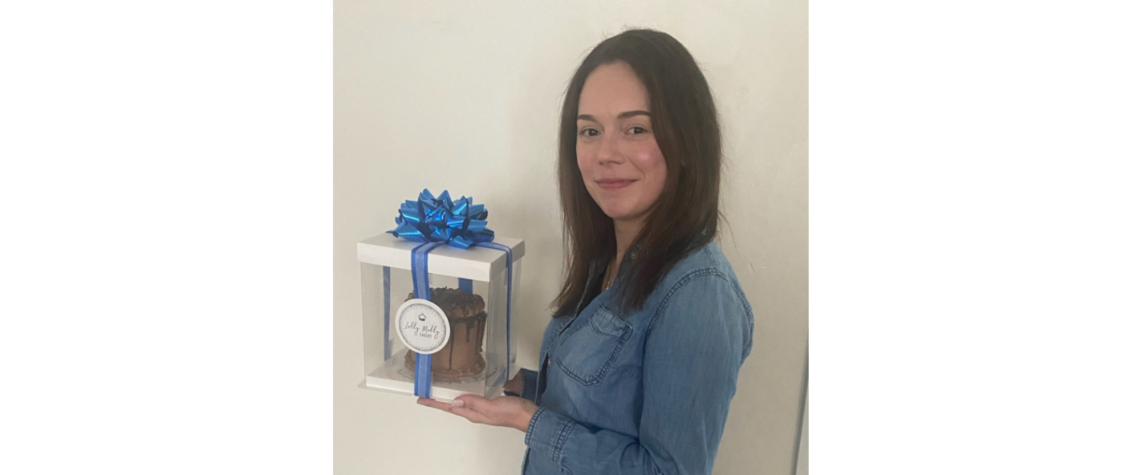 woman with long brown hair and a denim shirt holding a chocolate cake in a box with a blue ribbon