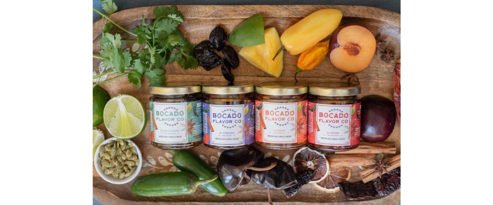 4 jars of chili crisp displayed on a wooden cutting board with fresh herbs, fruits and vegetables around the border