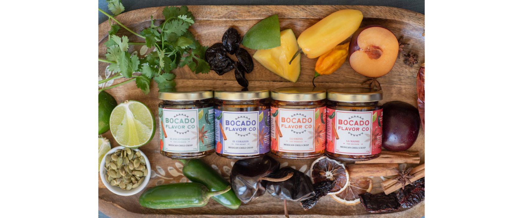 4 jars of chili crisp displayed on a wooden cutting board with fresh herbs, fruits and vegetables around the border